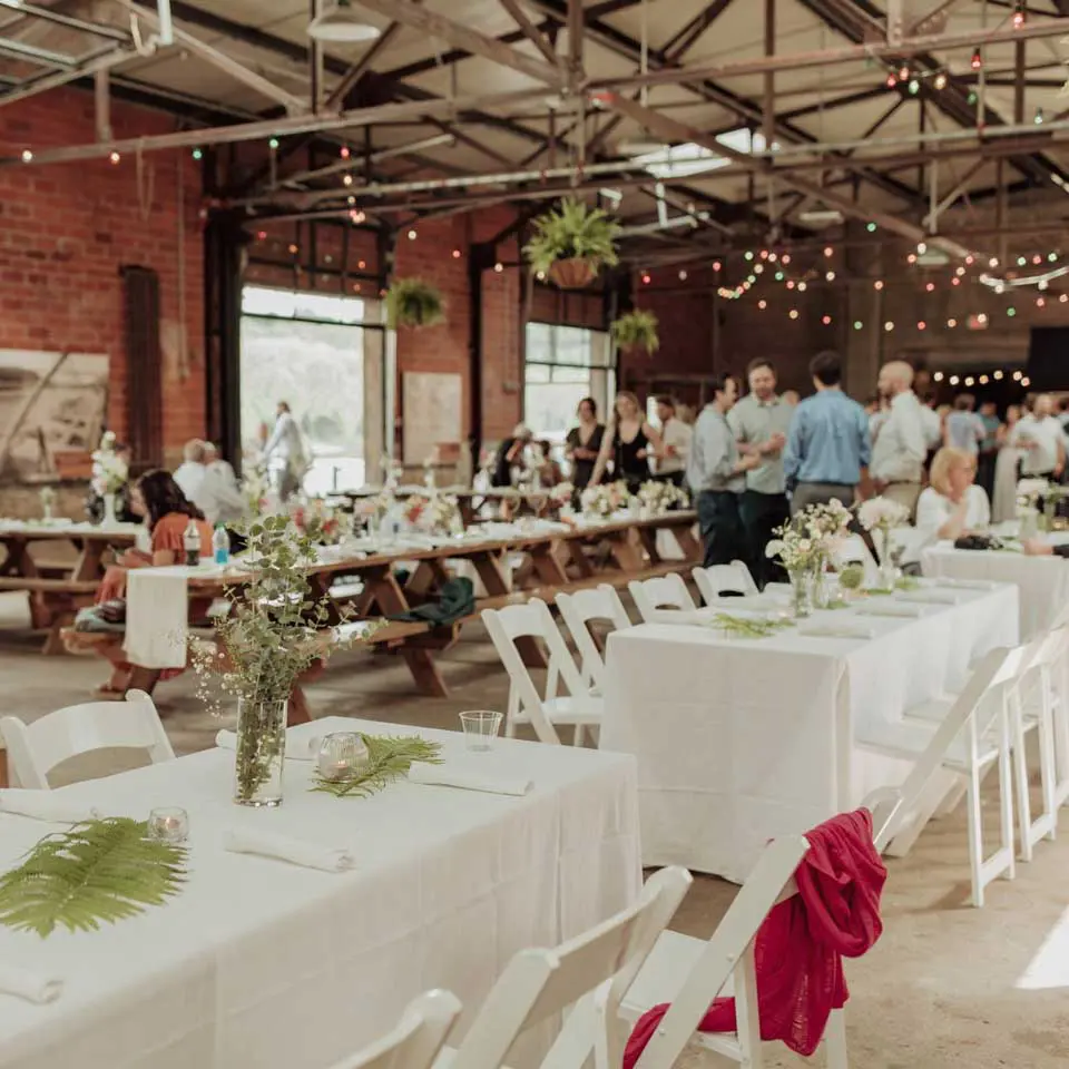 Wedding tables with white linen and white chairs set up while guest are in the background enjoying cocktail hour