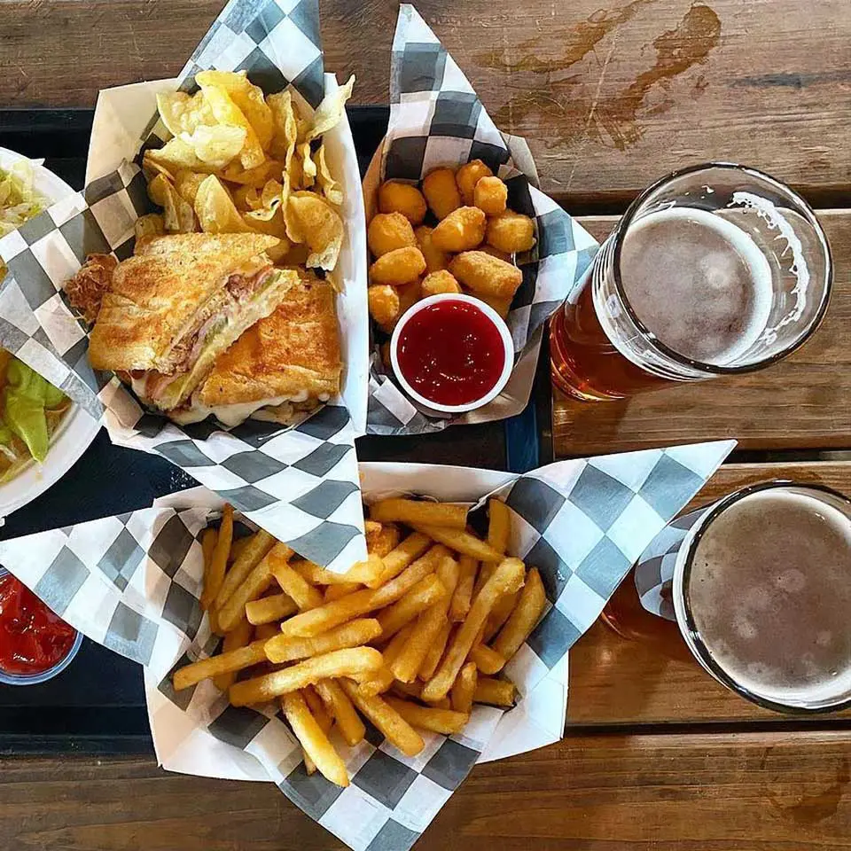 Overhead view of cubano sandwich, cheese curds and basket of fries with two glasses of beer on rustic wood table